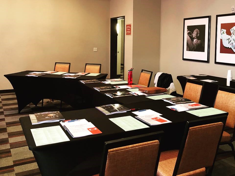 Conference room with tables arranged in a U-shape, covered with papers and brochures for an anger management training. Wall art of jazz musicians decorates the room.