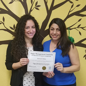 Two women smiling and holding a certificate in front of a painted tree mural. One woman, an anger management counselor, is presenting the certificate to the other.