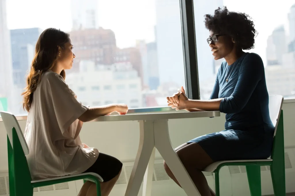 Two women sit across from each other at a table in a bright room, engaged in conversation with city buildings visible through the window behind them.