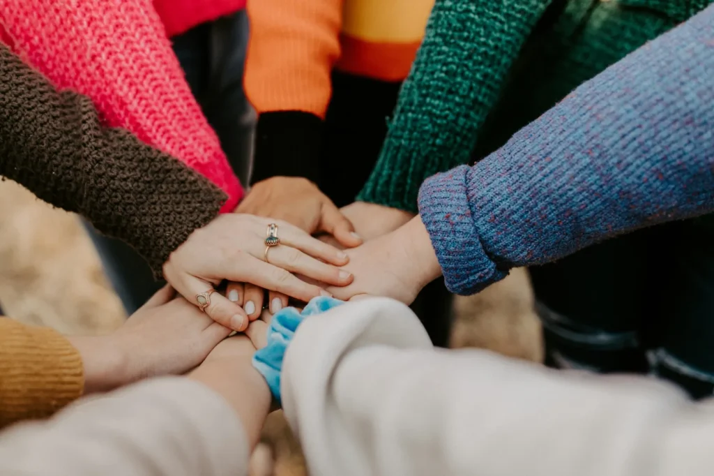 A group of people wearing colorful sweaters place their hands together in the center, forming a collaborative gesture.