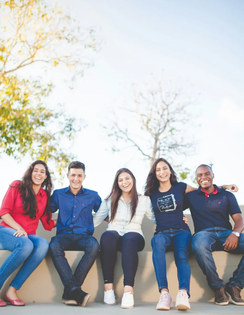 Five people sit outdoors on a ledge, smiling at the camera, with trees and blue sky in the background.